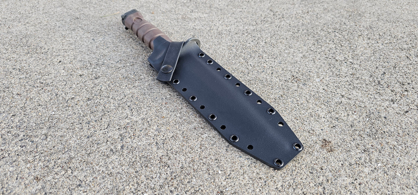 A black Kydex sheath for a OKC 
Marine 3S bayonet with a cross guard strap and optional belt attachment, lying on a textured surface.