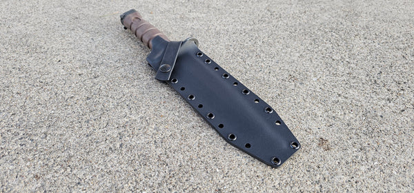 A black Kydex sheath for a OKC 
Marine 3S bayonet with a cross guard strap and optional belt attachment, lying on a textured surface.