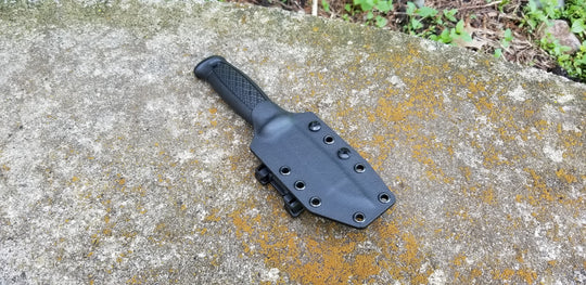 A black pancake style custom kydex sheath with polymer belt clip for a knife, placed on a textured surface.