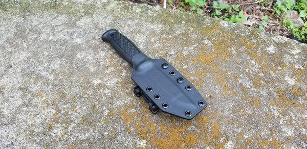 A black pancake style custom kydex sheath with polymer belt clip for a knife, placed on a textured surface.